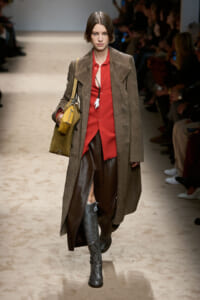 Female model walks a runway wearing a red blouse under a long brown coat, brown pants, snakeskin boots, and a mustard-yellow shoulder bag.