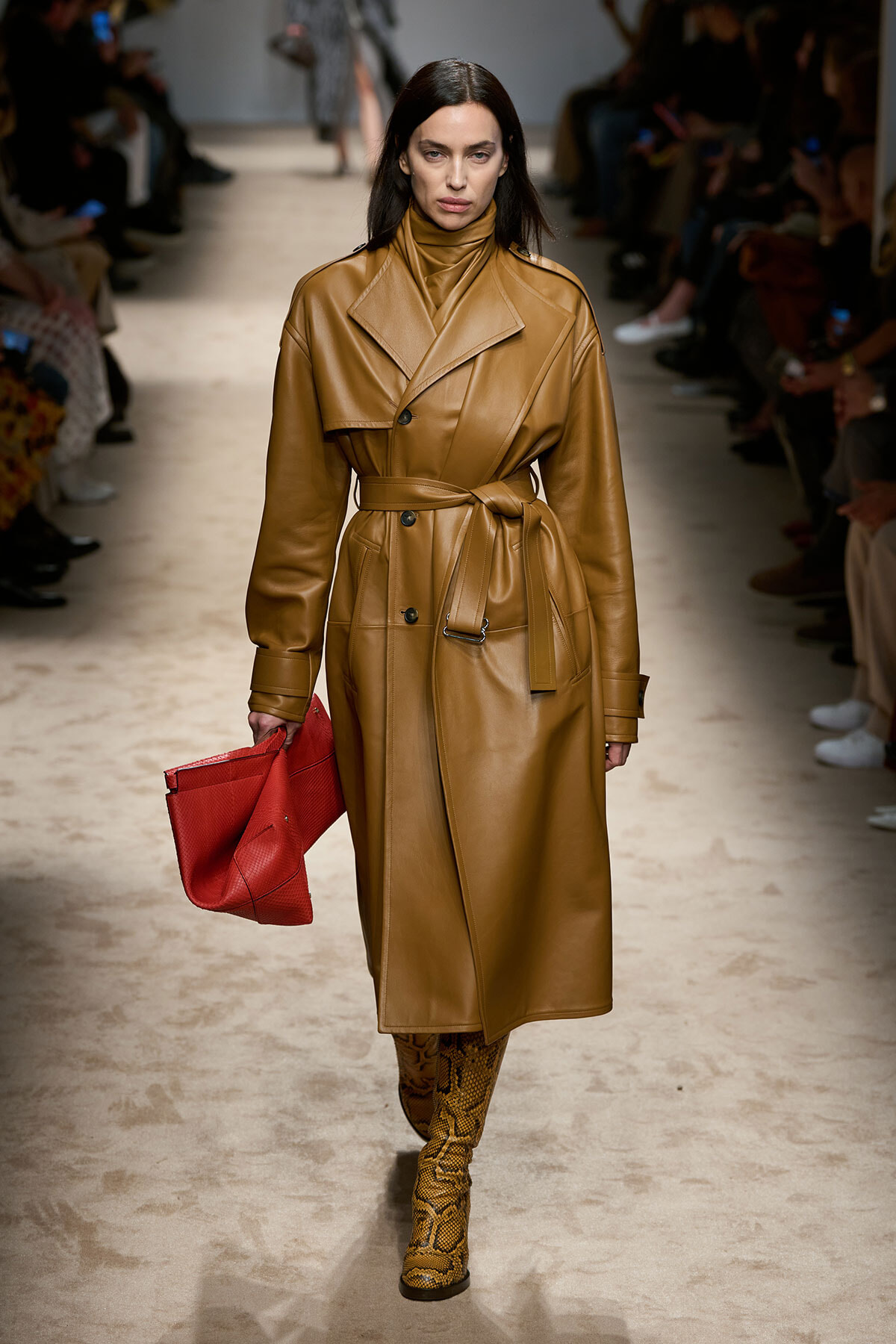 Model walking a runway in a tan leather trench coat with belt, holding a red clutch and wearing snakeskin boots.