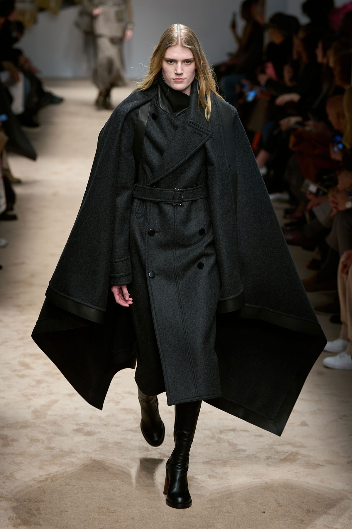 Model walking runway in a dark gray double-breasted cape coat with belt and tall boots.