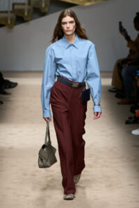 Model walking runway in a light blue button-down shirt tucked into burgundy wide-leg pants with a dark belt and small navy pouch at the waist, carrying a gray handbag and wearing snakeskin shoes