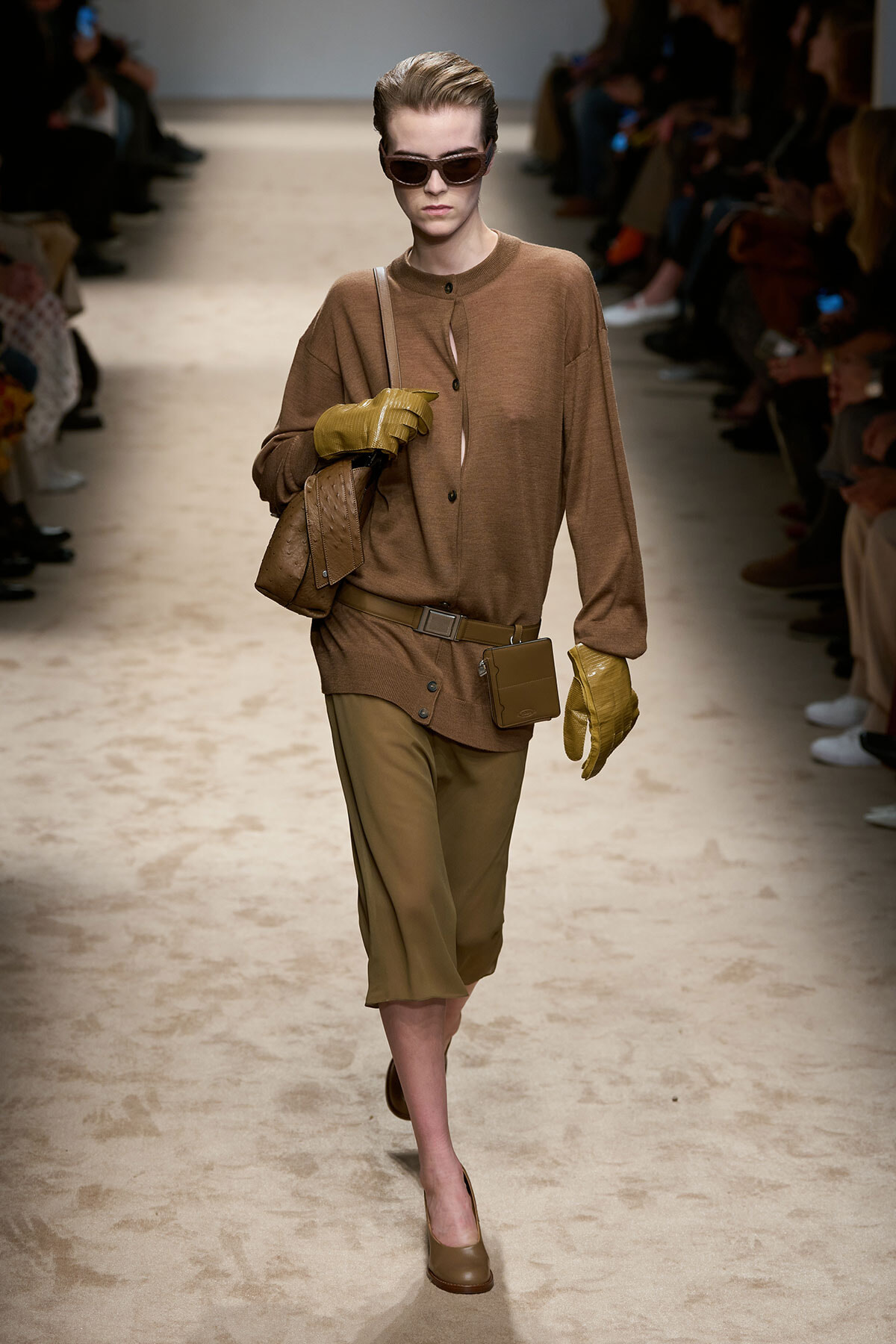 Model on a runway wearing a brown knit top, tan knee-length shorts, yellow gloves, tan crossbody bag, and a small belt pouch with dark sunglasses.