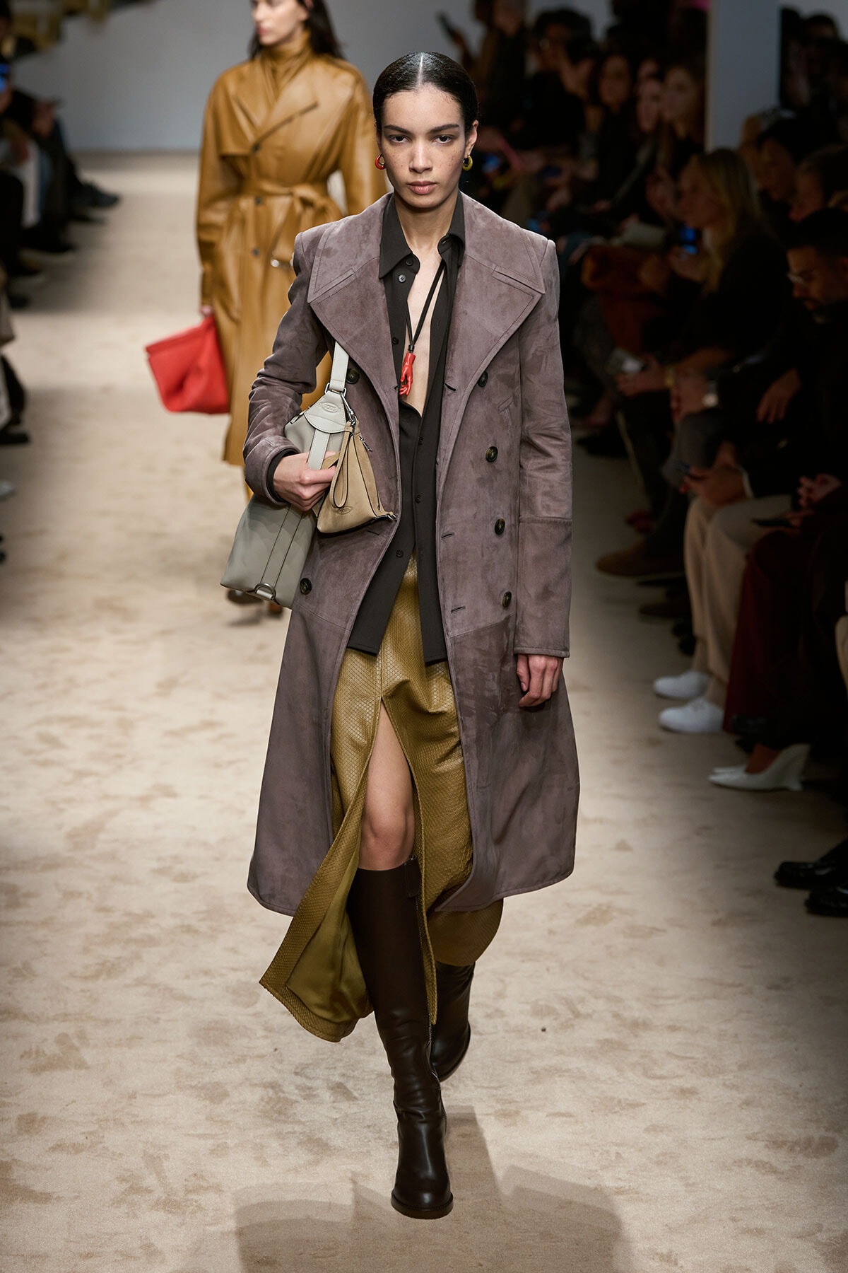 Runway model wearing a mauve long coat over a black blouse and gold slit skirt, knee-high boots, carrying a light gray and beige handbag.