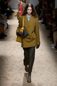 Model walks a runway in an oversized camel double-breasted blazer with a blue shirt, olive trousers, and tan ankle boots, carrying mustard crossbody bag and a dark blue handbag while wearing brown gloves.