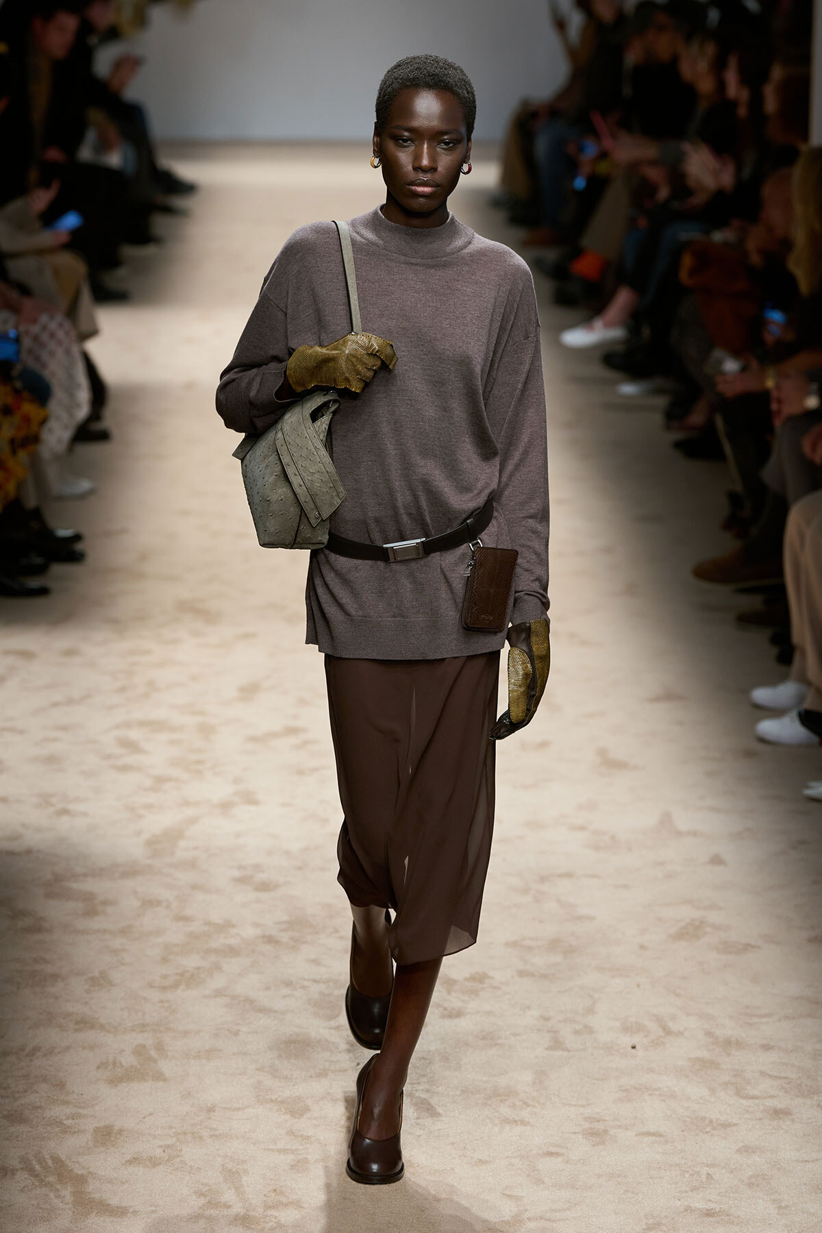 Model walks a runway in a taupe oversized sweater, brown draped skirt, brown heels, with a gray shoulder bag and gold gloves; audience in background.