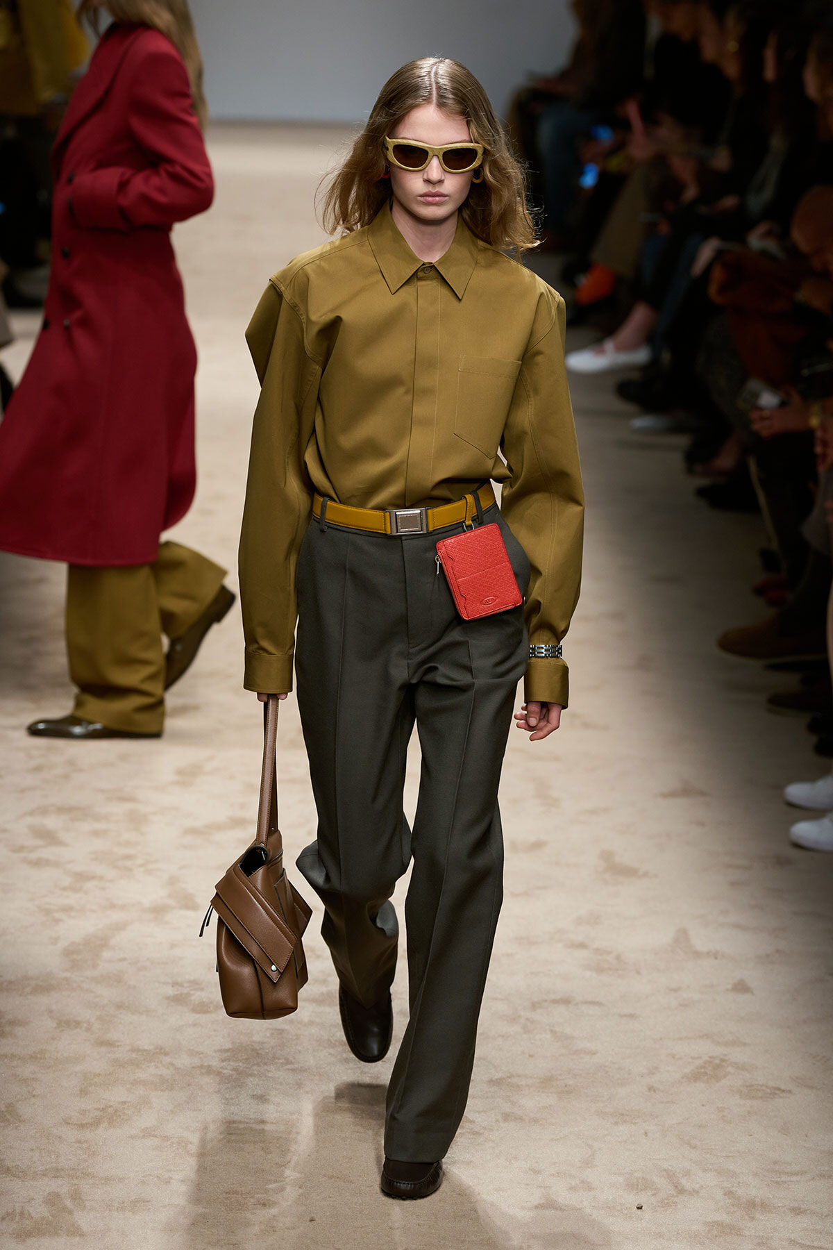 Runway model in an olive shirt with dark trousers, yellow belt, red cardholder, brown bag, and yellow sunglasses.