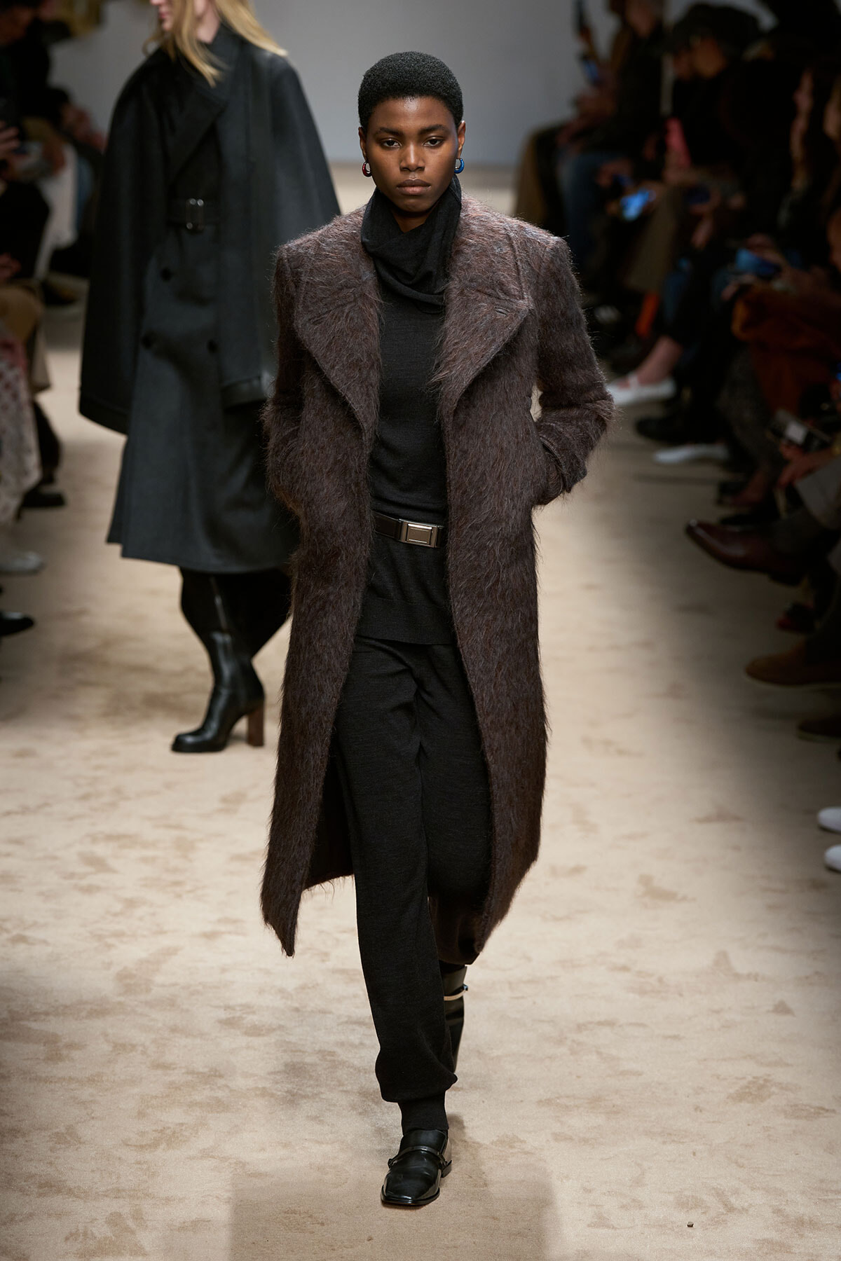 Model on a runway wearing a long, textured brown coat over a black turtleneck and black pants, with a black belt and loafers.
