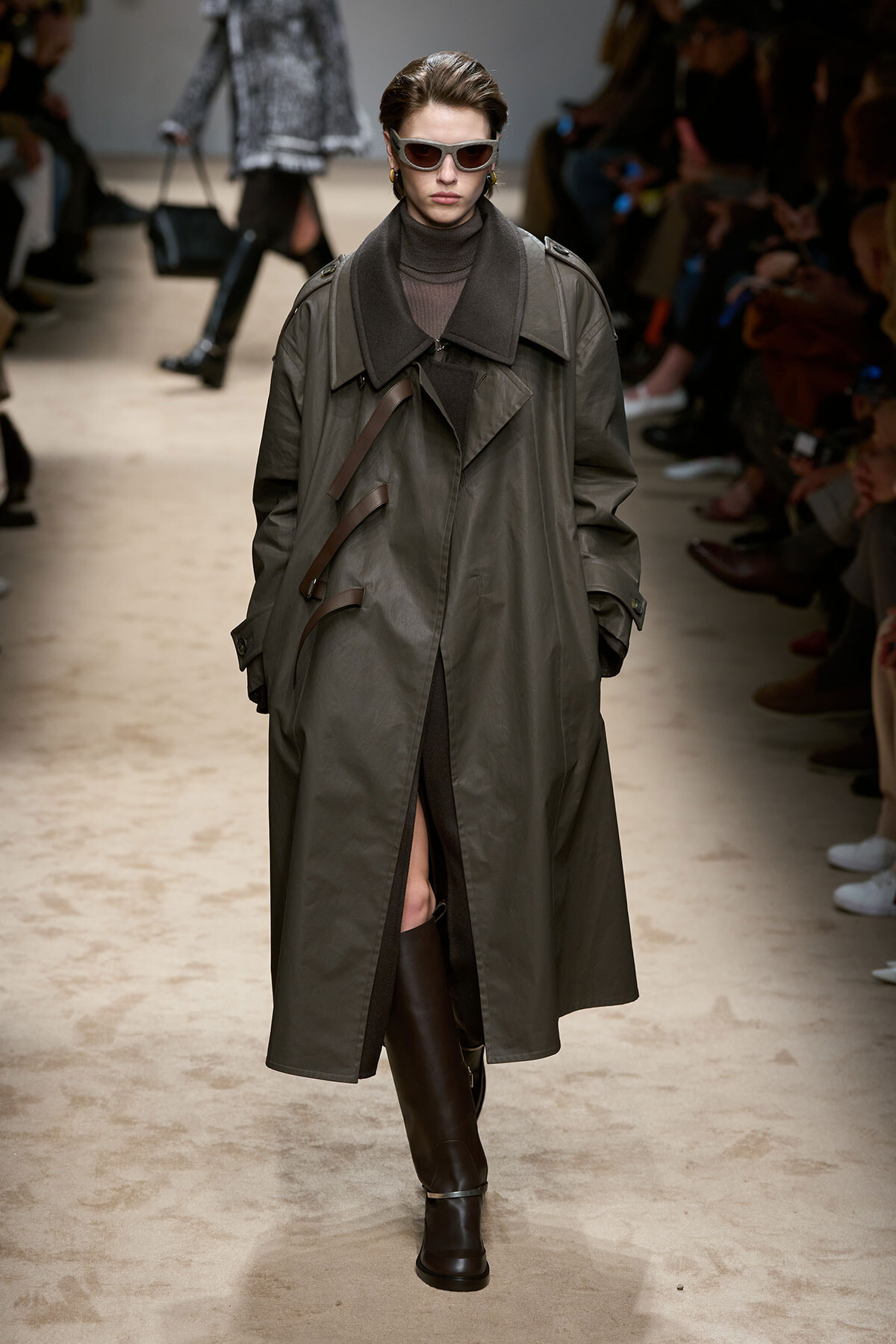Runway model in an oversized gray trench coat with leather strap details, black turtleneck, knee-high boots, and sunglasses.