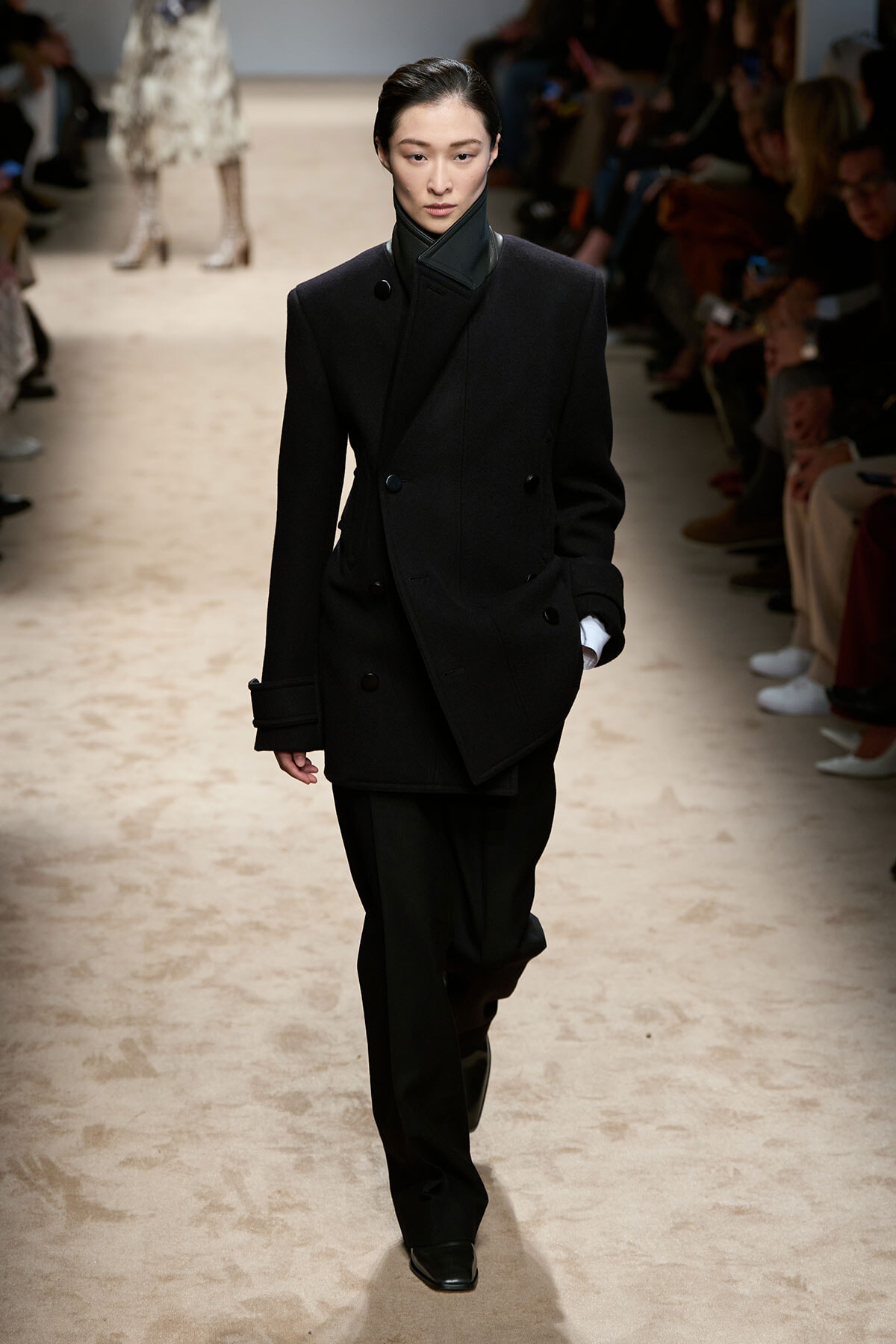 Model walks the runway in a black double-breasted oversized coat with a high collar and black trousers.