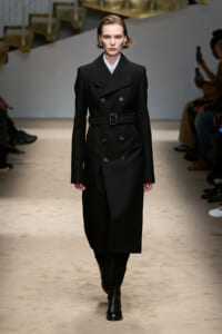 Model walking a fashion runway in a black double-breasted belted overcoat and black ankle boots.