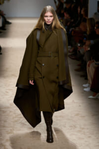 Model walks the runway wearing an oversized olive-green coat with a belt and dark leather accents, paired with knee-high boots.