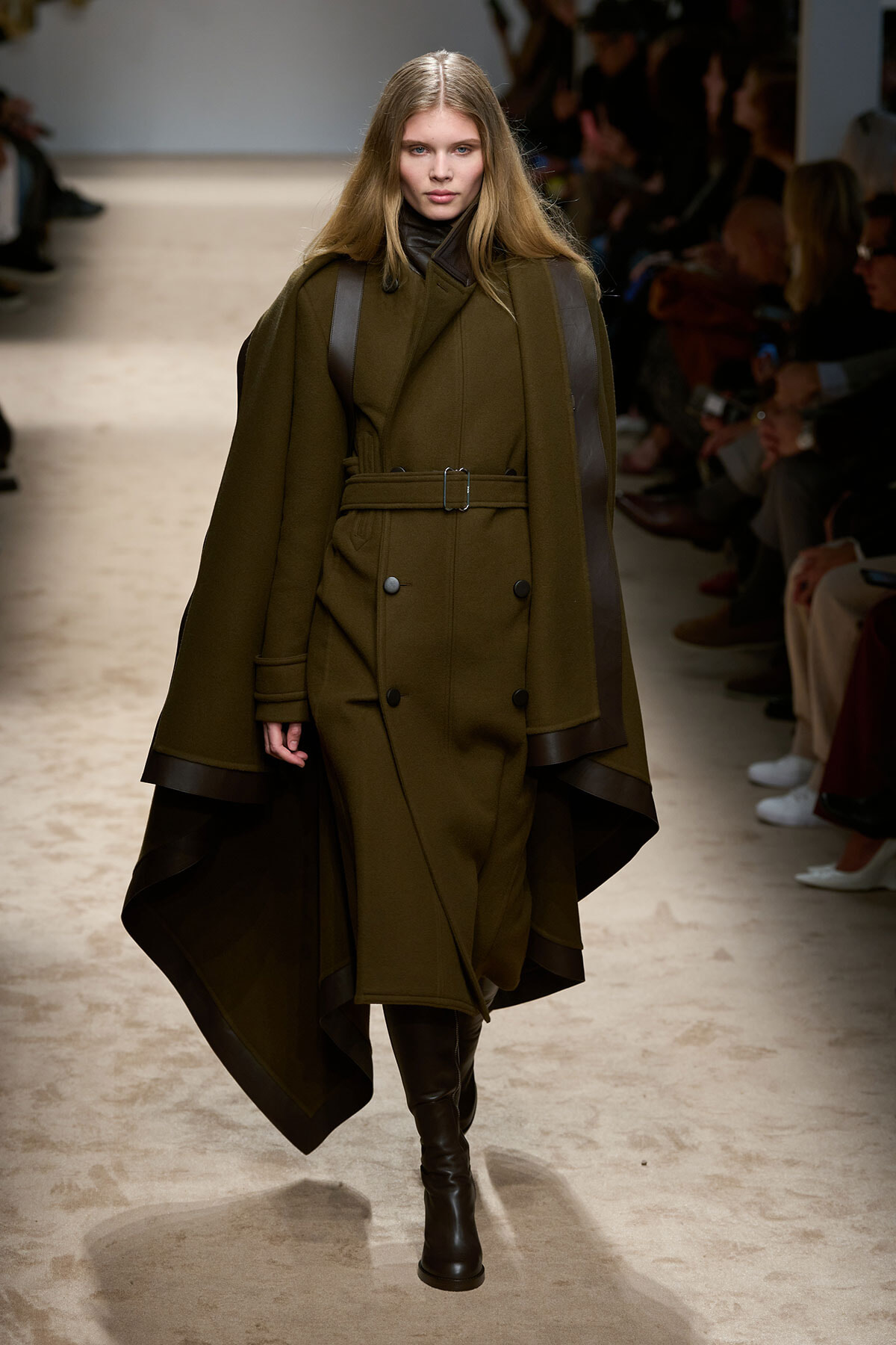 Model walks the runway wearing an oversized olive-green coat with a belt and dark leather accents, paired with knee-high boots.