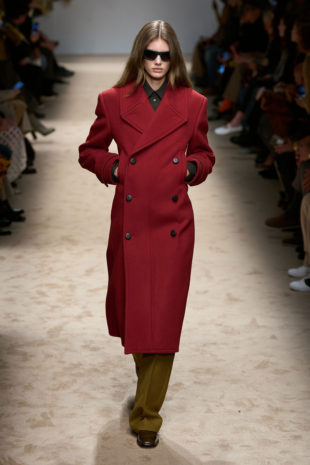 Model on a runway wearing a red double-breasted coat, black shirt, olive trousers, and dark sunglasses.