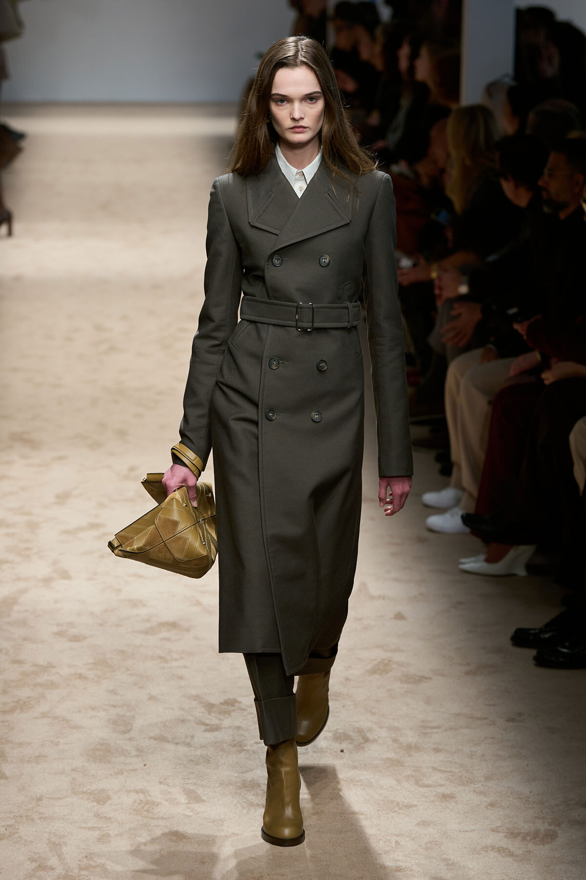 Model walking a runway in a dark gray double-breasted trench coat with a matching belt, paired with tan ankle boots and a tan geometric clutch bag