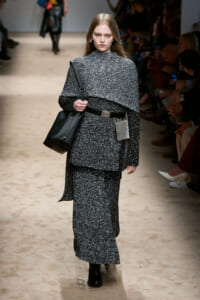Model walking a fashion show runway in a dark gray tweed layered outfit with a cape-like top, matching long skirt, black belt, and black knee-high boots, carrying a large black shoulder bag and a small tag hanging from the belt.