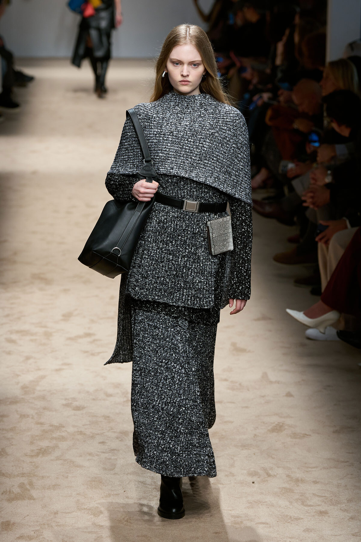 Model walking a fashion show runway in a dark gray tweed layered outfit with a cape-like top, matching long skirt, black belt, and black knee-high boots, carrying a large black shoulder bag and a small tag hanging from the belt.