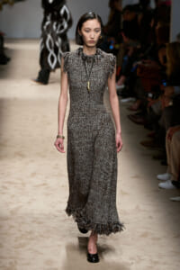 Model walks a runway wearing a floor-length textured grey tweed dress with fringe trim at sleeves and hem, accessorized with a long pendant necklace.
