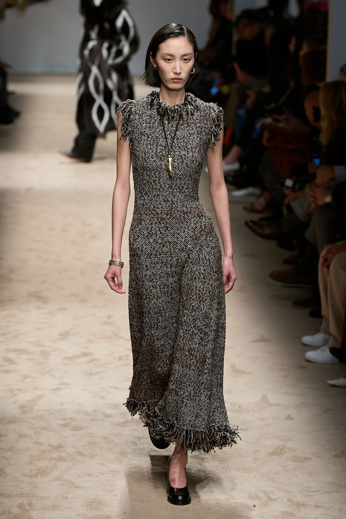 Model walks a runway wearing a floor-length textured grey tweed dress with fringe trim at sleeves and hem, accessorized with a long pendant necklace.