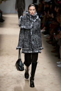 Model on a runway wearing a textured black-and-white fringed coat, carrying a black bag and wearing tall black boots.
