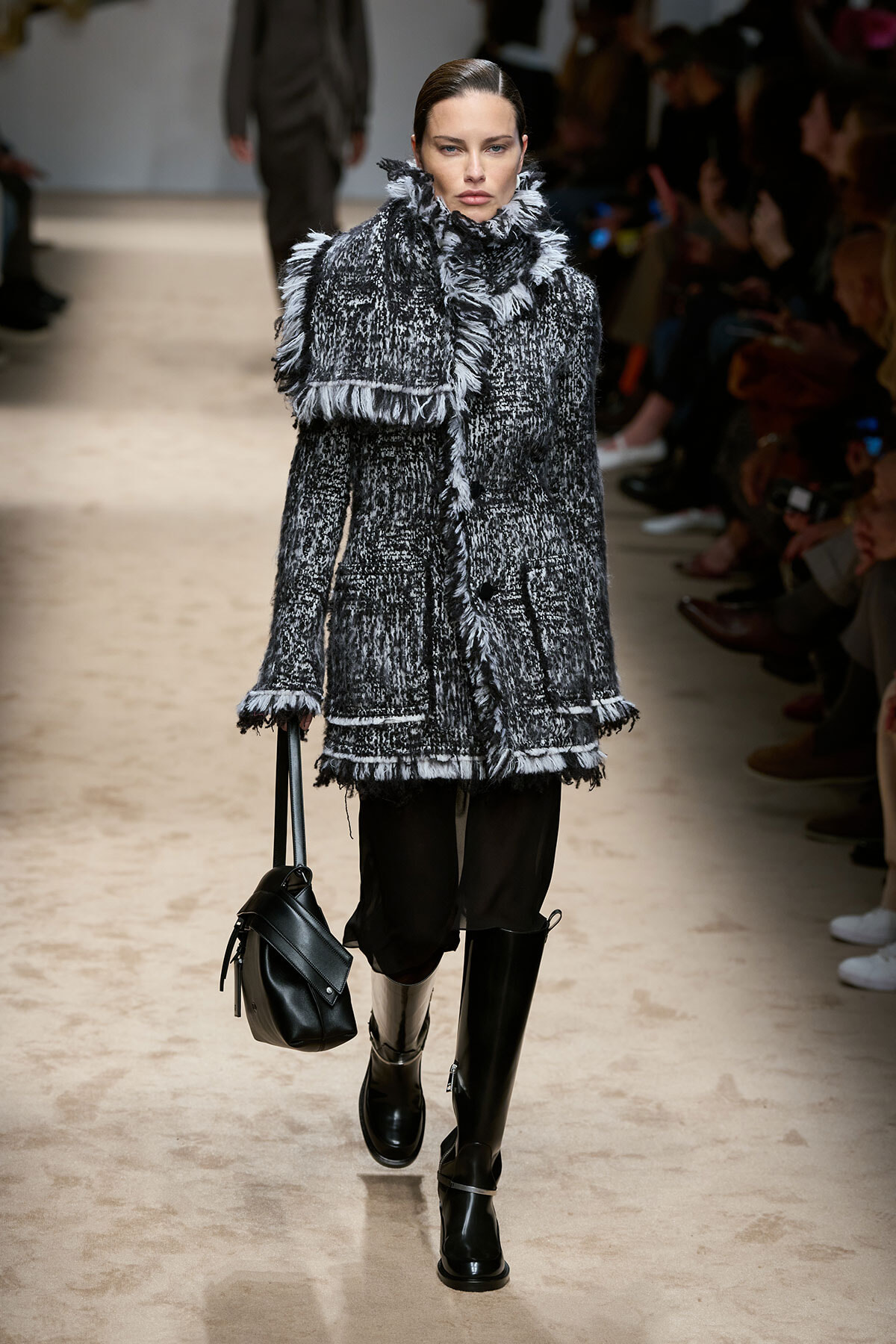Model on a runway wearing a textured black-and-white fringed coat, carrying a black bag and wearing tall black boots.