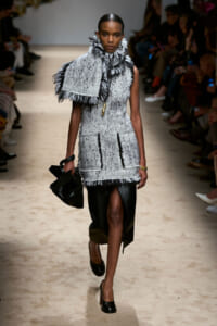 Model walks the runway wearing a gray tweed sleeveless dress with fringe trim and a large fringed scarf draped over her shoulder, paired with a black midi skirt and heels, holding a black sculptural bag nearby on the catwalk.