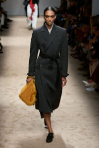 Female model walking a runway in a dark gray double-breasted belted coat, holding a yellow fur bag.