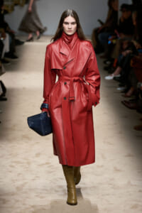 Model walks a runway in a bold red leather trench coat with belt, knee-high brown boots, and a dark blue textured clutch bag.