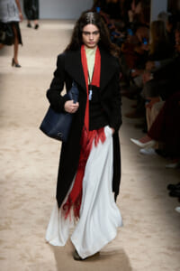 Model on a fashion runway wearing a long black coat over a pale yellow top, red fringe scarf, white draped skirt with red fringe, and a dark blue handbag.
