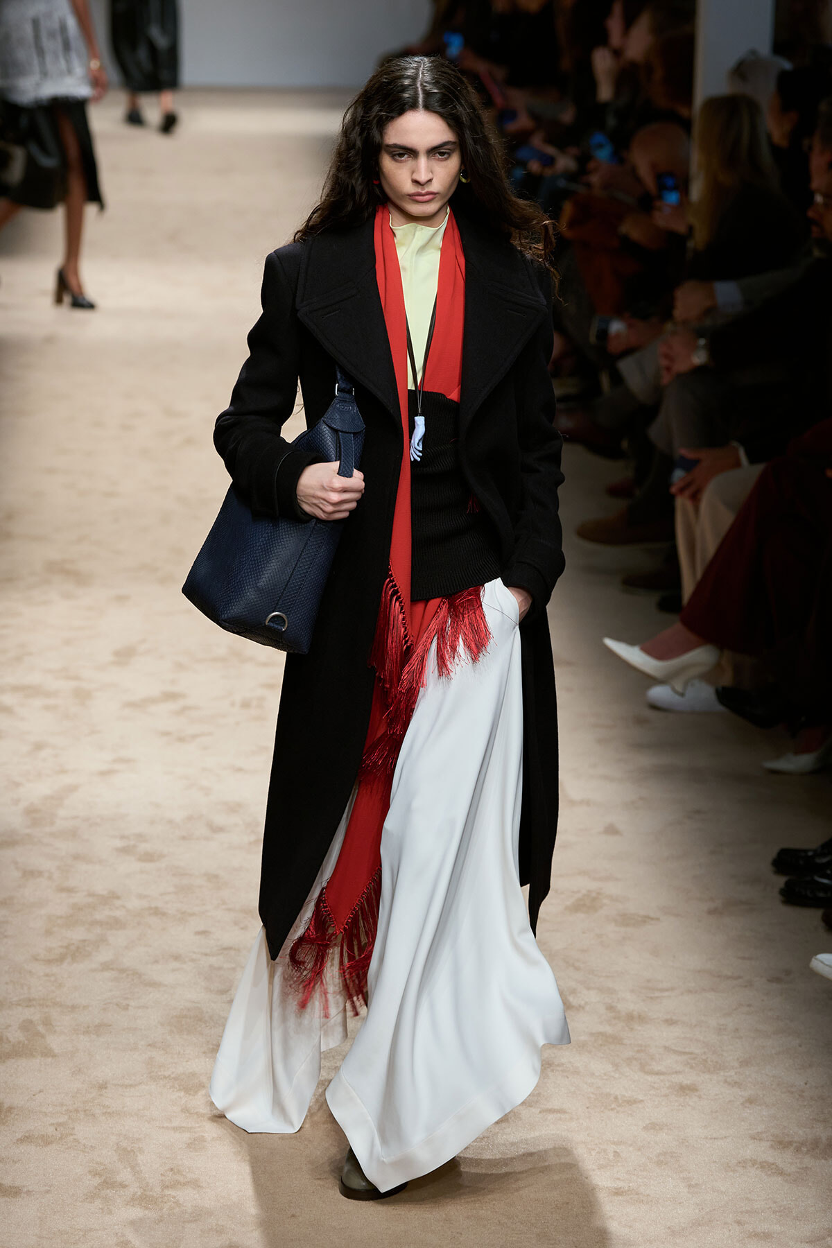 Model on a fashion runway wearing a long black coat over a pale yellow top, red fringe scarf, white draped skirt with red fringe, and a dark blue handbag.