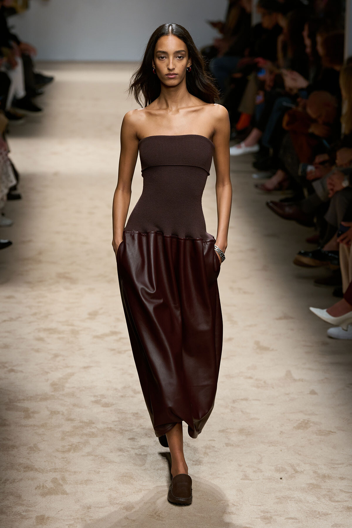 Model walks the runway in a strapless brown knit top and high-waisted burgundy leather pants with pockets.