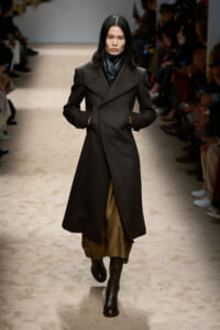 Model walks a runway in a long dark wool coat with wide lapels, brown wide-legged pants, and black ankle boots; audience seated along the sides.