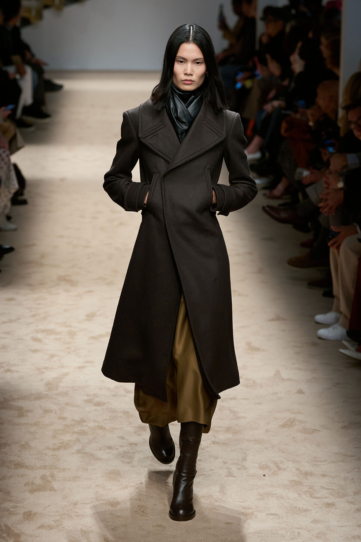 Model walks a runway in a long dark wool coat with wide lapels, brown wide-legged pants, and black ankle boots; audience seated along the sides.