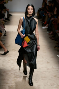 Model walking a runway in a black leather sleeveless dress with knee-high boots, carrying blue bag and colorful pouches at the waist.