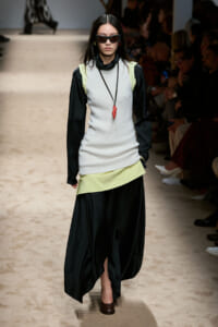 Model walking a runway in a layered outfit: light gray sleeveless top with lime accents over a black blouse and a long black skirt, wearing sunglasses and a red pendant necklace.