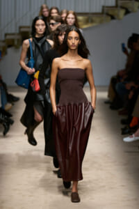 Model walks the runway in a strapless brown top and burgundy leather skirt; lineup of models behind her.