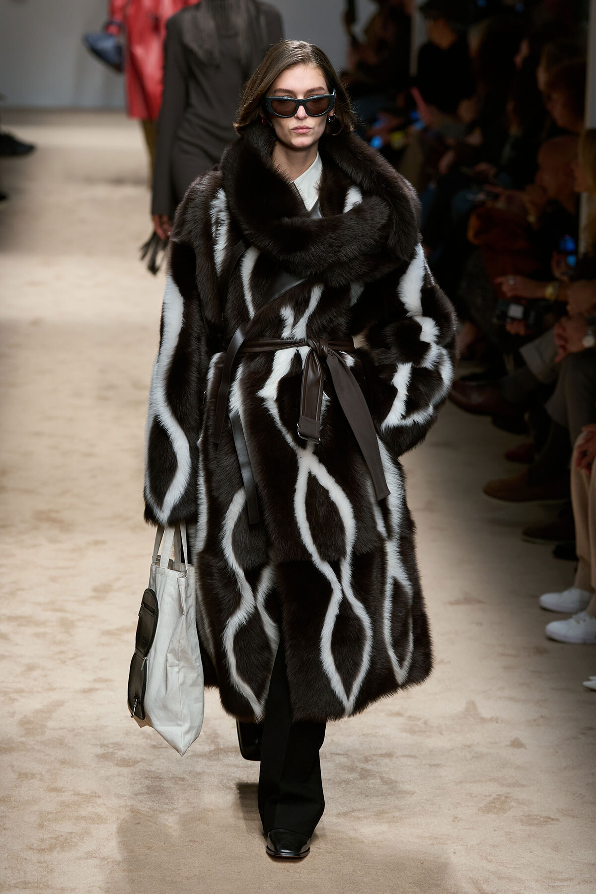 Model on a runway wearing a large black-and-white fur coat with a brown belt, sunglasses, and a light tote bag ready for the next look.