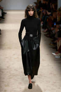 Model walking runway in black turtleneck and full black leather skirt with belt and small pouch at the waist.