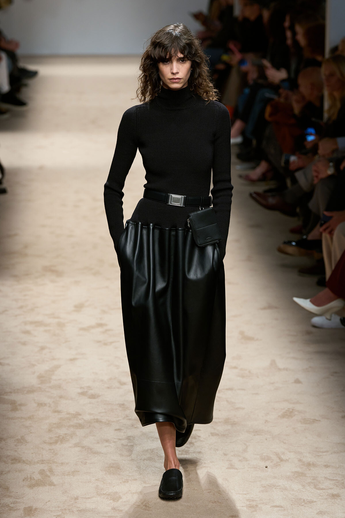 Model walking runway in black turtleneck and full black leather skirt with belt and small pouch at the waist.