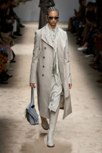 Runway model in a long beige trench with fringe scarf, cream boots, and gray handbag strides down a fashion show aisle wearing sunglasses.