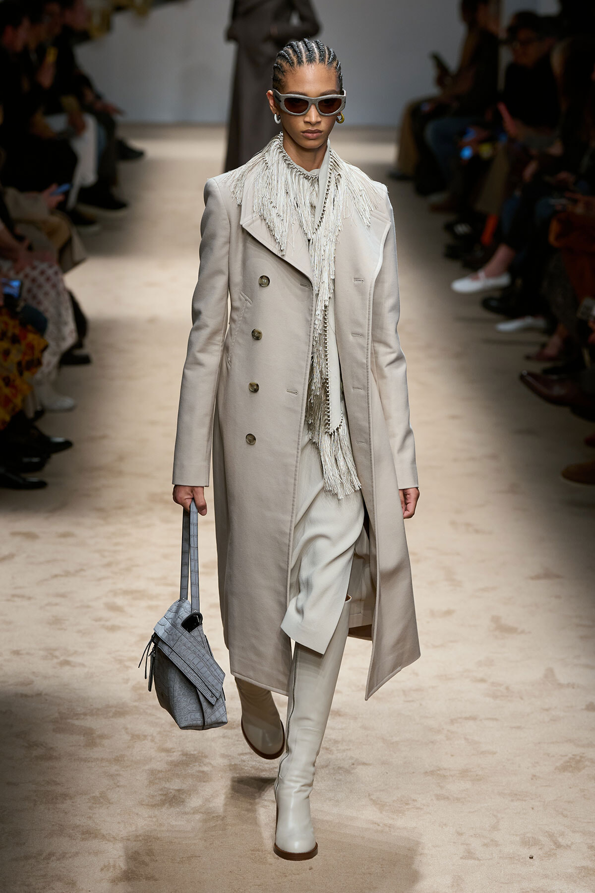 Runway model in a long beige trench with fringe scarf, cream boots, and gray handbag strides down a fashion show aisle wearing sunglasses.