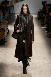Model walking a runway in a long dark brown leather trench and black outfit, carrying a black bag and wearing tall boots.