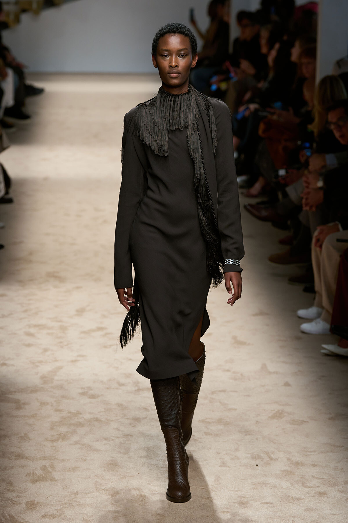 Model walks down a runway in a long black dress with a fringed scarf, pairing knee-high brown boots at a fashion show.