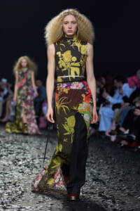 Model walks a runway in a black floral-print sleeveless top with matching wide-legged pants, surrounded by a seated audience and confetti on the floor.
