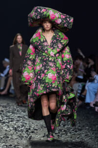 Model walking a fashion runway in a oversized floral-print coat with matching hat and bag, black background stage.