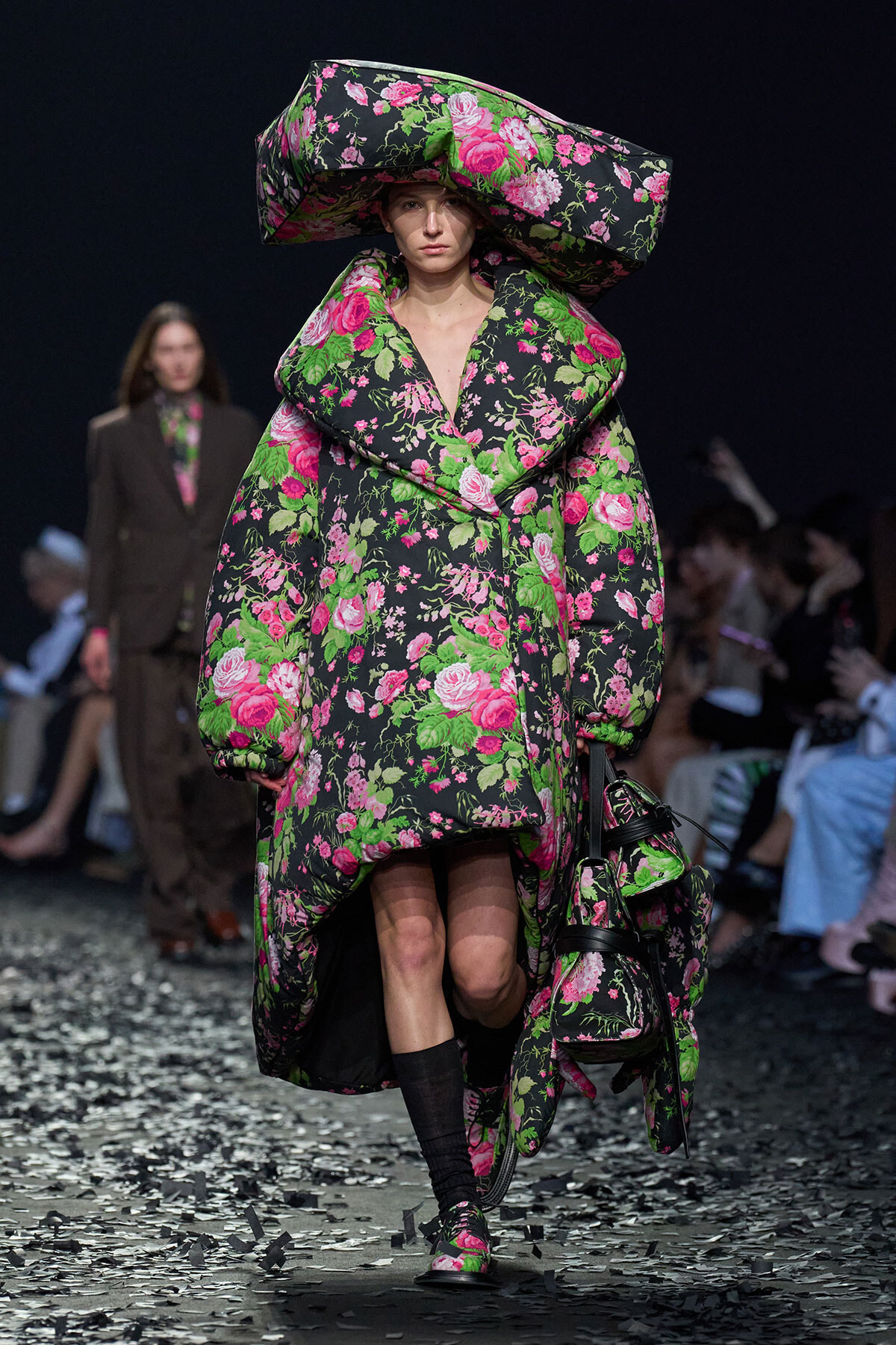 Model walking a fashion runway in a oversized floral-print coat with matching hat and bag, black background stage.