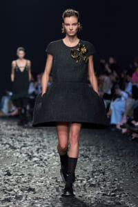 Model on a runway wearing a black textured short dress with a gold floral brooch on the bodice, confetti on the floor.