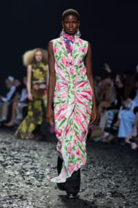 Model walks the runway in a sleeveless pink and green floral dress with a draped skirt, dark backdrop and audience nearby.