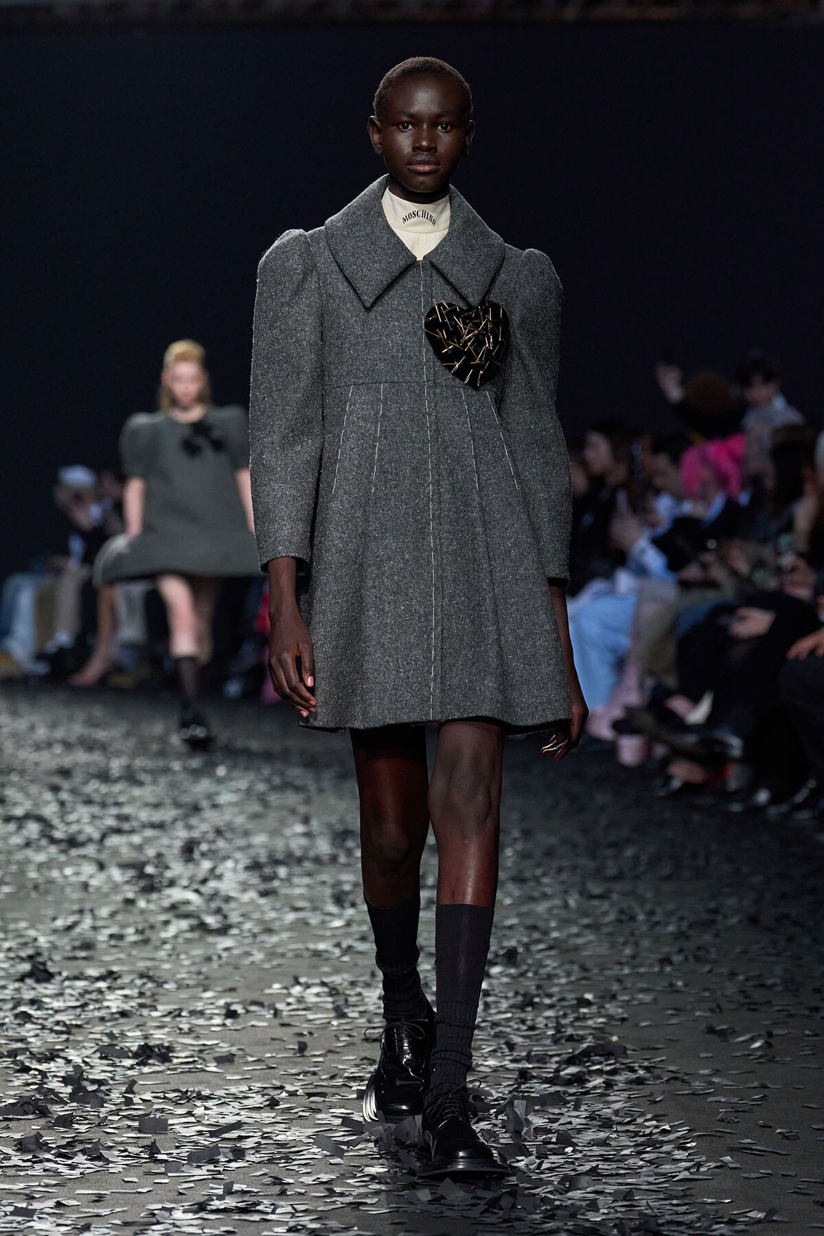 Model walks a grey wool coat with large lapels and a heart-shaped brooch on the chest, on a fashion show runway with confetti.