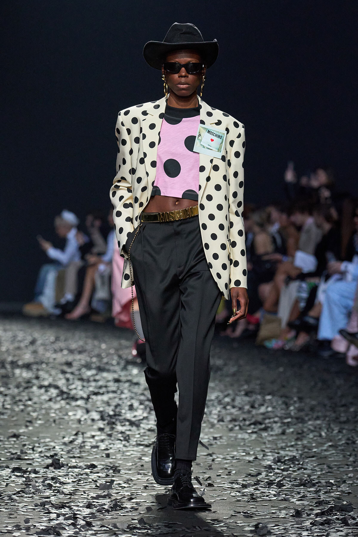 Runway model in a cream polka-dot blazer over a pink cropped top, black trousers, belt, hat and sunglasses with a crowd in the background.