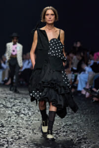 Model walks the runway in a black asymmetrical dress with white polka dots and layered ruffles, confetti on the floor.