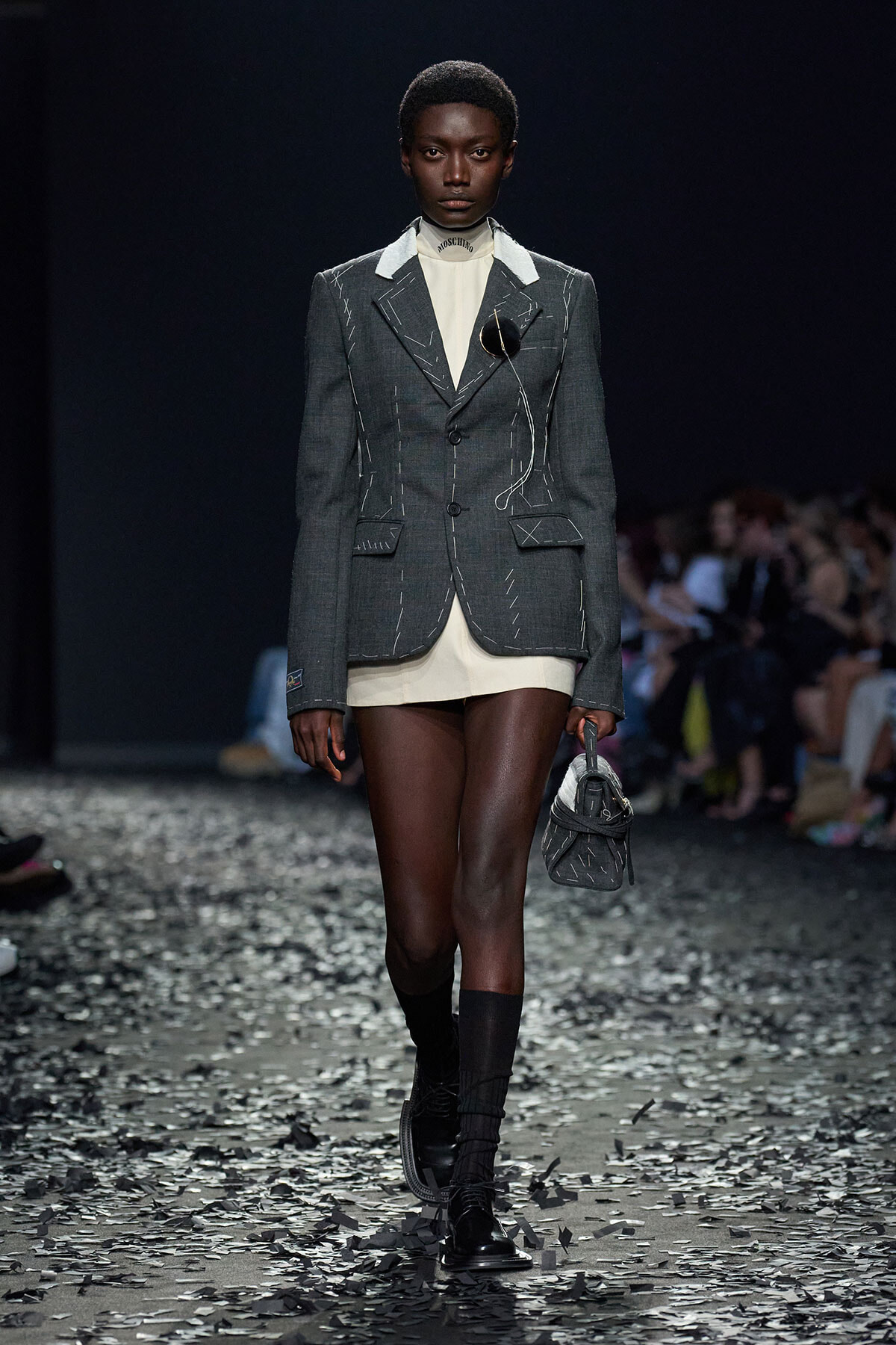 Model walking a runway in a gray stitched blazer, cream top, and matching shorts, carrying a small gray handbag amid confetti on the floor.